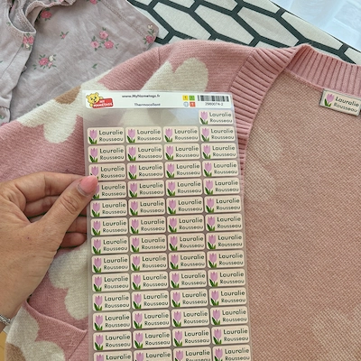 Main tenant une planche d’étiquettes thermocollantes devant un gilet rose étiqueté